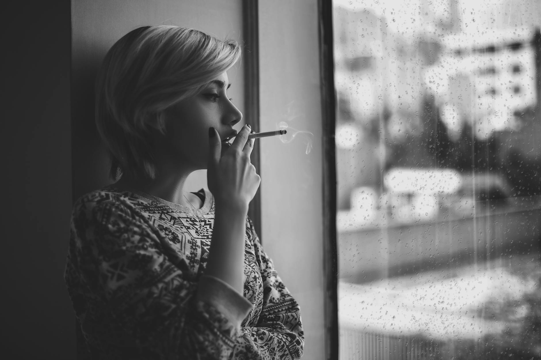 melancholic woman smoking cigarette near window during rain